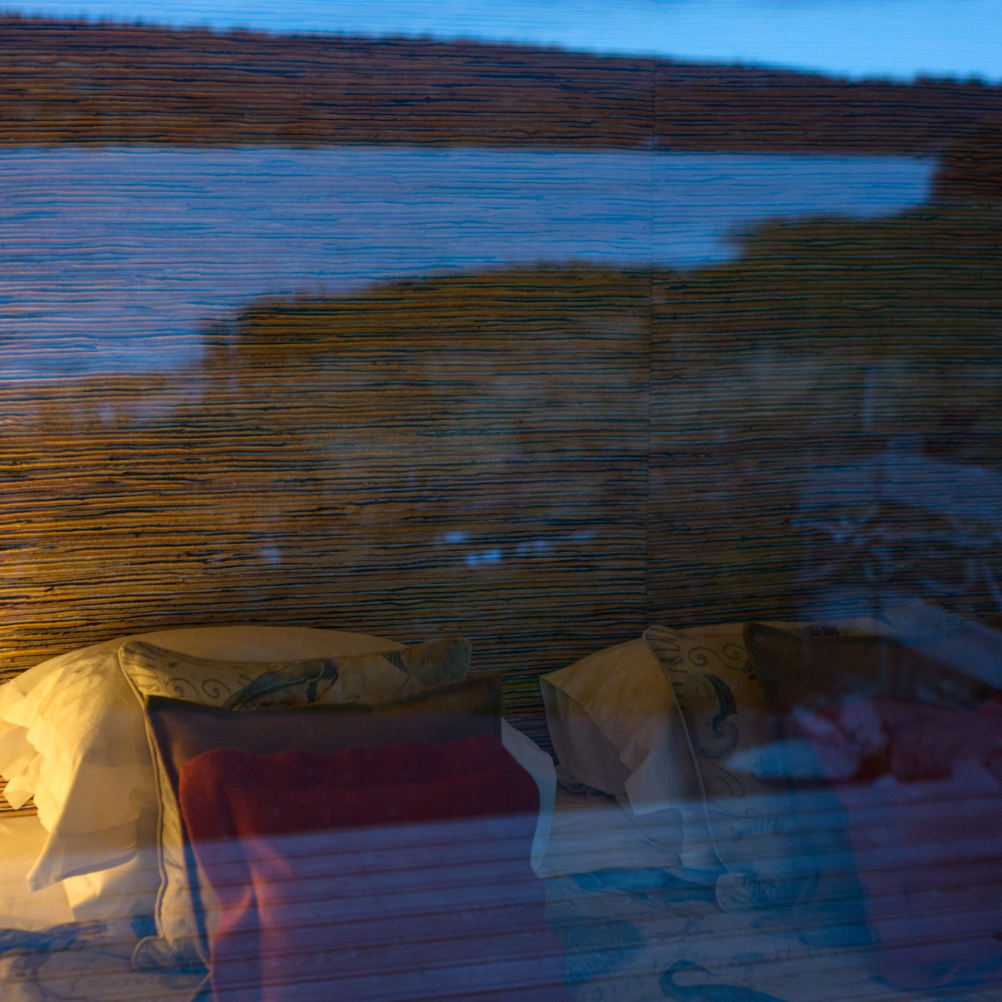 Un lit aux coussins clairs vu à travers une vitre, sur lequel se superpose le reflet du lac et des rives au crépuscule, dans une lumière bleue et froide.
