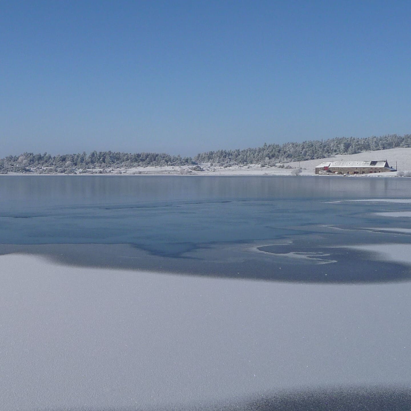 Le lac du Pêcher entièrement gelé sous un ciel bleu vif, l'écolodge apparaît au loin sur la rive enneigée, fondu dans le paysage.