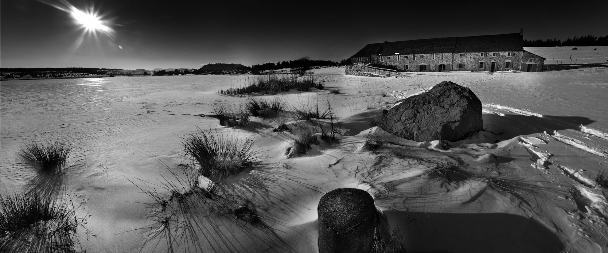 L’écolodge en pierre au bord du lac du Pêcher gelé, sous un soleil bas d’hiver, la neige balayée au premier plan.